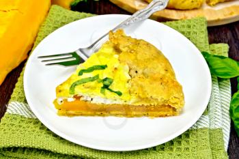 One piece of the pie of pumpkin, salty feta cheese, eggs, cream and herbs in a plate on a napkin, basil on a wooden boards background