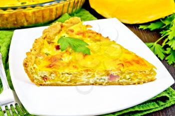 Piece of pie kish with pumpkin and bacon, filled with milk with eggs and cheese in a plate on a napkin, parsley on a background of a dark wooden board