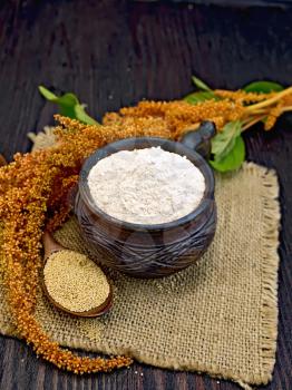 Flour amaranth in a clay cup, a spoon with grain, brown flower with green leaves on a napkin from a sacking on a background of wooden boards
