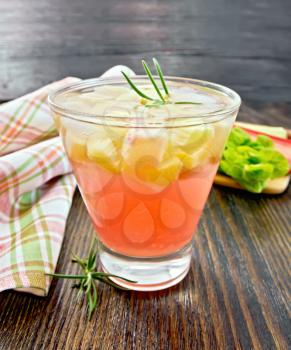 Lemonade with rhubarb and rosemary in a glass, stems and a leaf of a vegetable, napkin on a wooden plank background