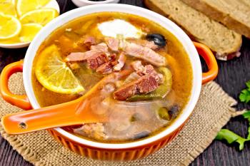 Soup saltwort with lemon, meat, pickles, tomato sauce olives, spoon in a bowl on a napkin of burlap on the background of a wooden board