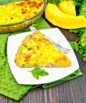 Piece of pie kish with pumpkin and bacon, filled with milk with eggs and cheese in a plate on a napkin, parsley on a wooden board background