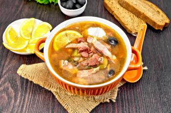 Soup saltwort with lemon, meat, pickles, tomato sauce olives in a bowl on a sacking, bread, parsley and spoon on a wooden board background
