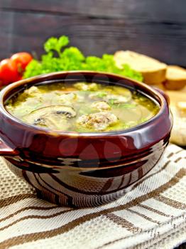 Soup with meatballs, noodles and champignon in a clay bowl on a towel, parsley, tomatoes, mushrooms and bread on dark background wooden plank