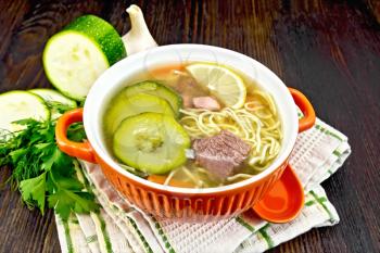 Soup with zucchini, beef, ham, lemon and noodles in a bowl, parsley and dill on a kitchen towel against a dark wooden board
