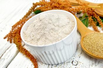 Amaranth flour in white bowl, spoons with grain, brown flower with leaves on the background of wooden boards