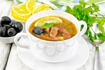 Soup saltwort with lemon, meat, pickles, tomato sauce and olives in a white bowl, towel, parsley on a light wooden board background