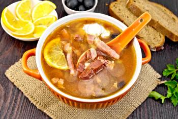 Soup saltwort with lemon, meat, pickles, tomato sauce olives, spoon in a bowl on a napkin of burlap on the background of a dark wooden board