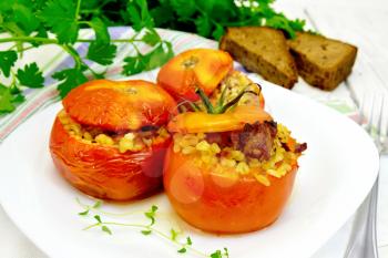 Tomatoes stuffed with meat and steamed wheat bulgur, a sprig of thyme in a plate, napkin, fork, bread and parsley on the background light wooden boards