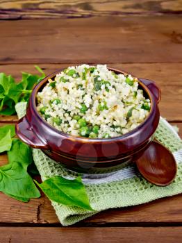 Couscous with spinach and green peas in a clay bowl on a towel, basil and spoon on a wooden plank background