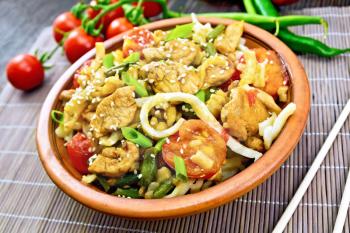 Thai noodles wok with chicken meat, tomatoes, soy sauce and green beans sprinkled with sesame seeds in a clay dish on a bamboo napkin on a wooden board background