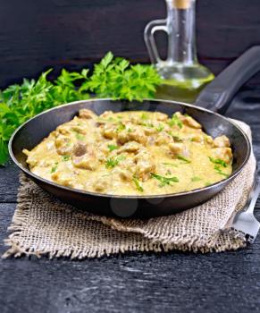 Meat stewed with cream in an old frying pan on burlap, parsley, fork and vegetable oil on a black wooden board background