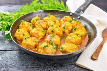 Meatballs with tomato sauce in a frying pan, parsley, dill, napkin and spoon on a black wooden board background