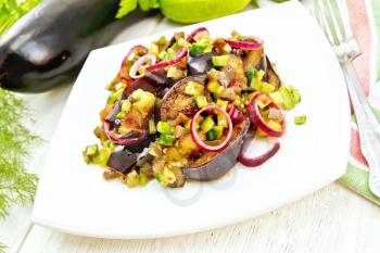 Salad of fried eggplant, fresh and pickled cucumber with red onion, seasoned with vegetable oil and spicy sauce in a plate, towel, fork and dill on a light wooden board background