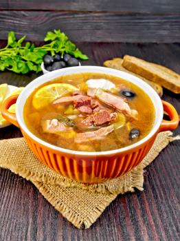 Soup saltwort with lemon, meat, pickles, tomato sauce and olives in a sackcloth, bread, parsley and spoon on a background of a dark wooden board