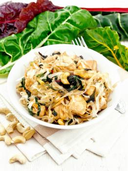 Rice noodles with leafy beet, chicken breast, cashew nuts and soy sauce in a bowl on a towel against a light wooden board