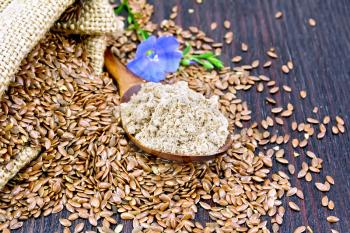 Flaxseed flour in a spoon, seeds in a bag and on a table, blue linen flower on a background of a dark wooden board