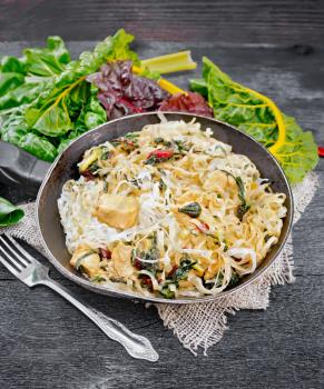 Rice noodles with leafy beet, chicken breast meat, cashew nuts and soy sauce in a frying pan on a sacking on the background of a black wooden board