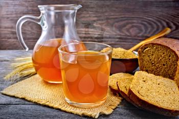 Kvass in glassful and glass jug on burlap, malt in a bowl, rye bread on wooden plank background