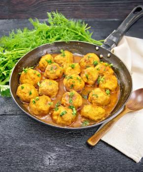 Meatballs with tomato sauce in a frying pan with parsley greens, dill, a dish towel and a spoon on the background of a wooden board