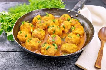 Meatballs with tomato sauce in a frying pan with parsley greens, dill, a dish towel and a spoon on the background of a black wooden board