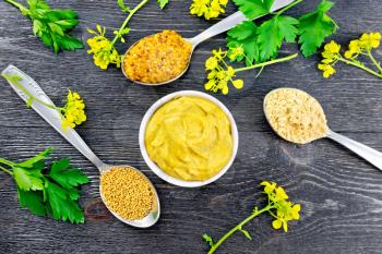 Mustard granular, powder, seeds in spoons and mustard sauce in a bowl, yellow flowers and parsley on the background of a wooden board from above