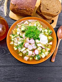 Cold soup okroshka from sausage, potatoes, eggs, radish, cucumber, greens and kvass in a bowl, bread and jug with drink on background of dark wooden board from above