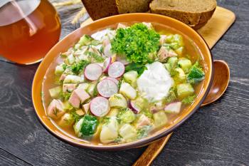 Cold soup okroshka from sausage, potato, egg, radish, cucumber, greens and kvass in a bowl, spoon, bread and jug with drink on the background of wooden board