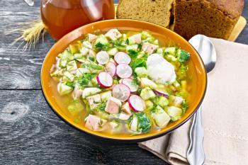 Cold soup okroshka from sausage, potatoes, eggs, radish, cucumber, greens and kvass in a bowl, kitchen towel, bread and jug with drink on background of a dark wooden board