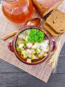 Cold soup okroshka from sausage, potatoes, eggs, radish, cucumber, greens and drink of kvass in a clay bowl, bread on napkin on wooden board background from above