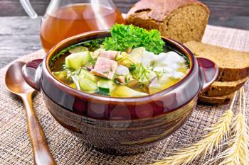 Cold soup okroshka from sausage, potato, egg, radish, cucumber, greens and drink of kvass in a clay bowl, bread on wicker napkin on background of dark wooden board
