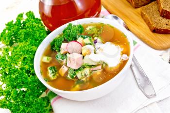 Cold soup okroshka from sausage, potatoes, eggs, radish, cucumber, greens and kvass in a white bowl on kitchen towel, bread and jug with drink on the background of light wooden board