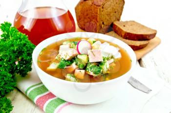Cold soup okroshka from sausage, potato, egg, radish, cucumber, greens and kvass in a white bowl on napkin, bread and jug with drink on the background of wooden board
