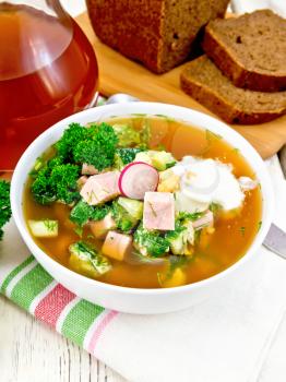Cold soup okroshka from sausage, potato, egg, radish, cucumber, greens and kvass in a white bowl on kitchen towel, bread and jug with drink on the background of wooden board