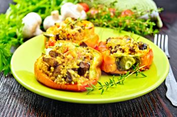 Pepper sweet, stuffed with mushrooms, tomatoes, couscous and cheese in a green plate on napkin, a fork, parsley against the background of dark wooden board