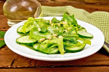 Salad from spinach, fresh cucumbers, rukkola salad, cedar nuts and spring onions, seasoned with vegetable oil on a plate, napkin and fork on a brown board background