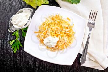 Salad of fresh carrots, kohlrabi cabbage with sour cream in a plate, napkin and fork on a dark wooden board background on top