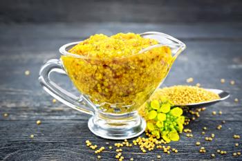 Mustard Dijon sauce in a glass sauceboat, yellow flowers and mustard seeds in a spoon on a black wooden board background