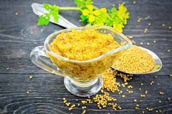 Mustard Dijon sauce in a glass sauceboat, yellow flowers and mustard seeds in a spoon on a wooden plank background