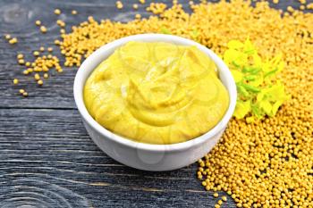 Mustard sauce in a white bowl, mustard flower and seeds on a dark wooden board background
