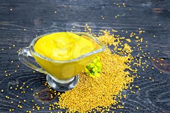 Mustard sauce in a glass sauceboat, seeds and flowers of mustard on a background of a dark wooden board