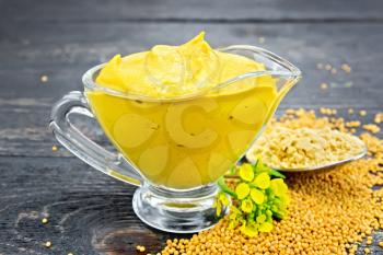 Mustard sauce in a glass sauceboat, seeds, flowers and mustard powder in a spoon on a black wooden board background