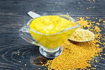 Mustard sauce in a glass sauceboat, seeds and mustard powder in a spoon on a black wooden board background
