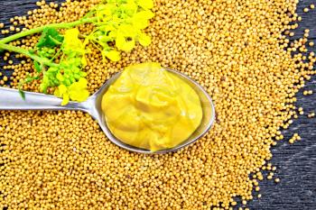 Mustard sauce in a metal spoon with a yellow flower on mustard seeds against the background of a wooden board from above