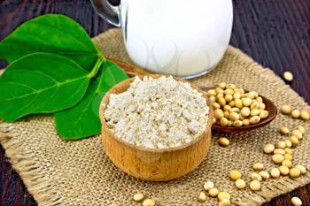 Soy flour in the bowl, soybeans on a napkin of burlap, milk in a jug, soya leaf against a dark wooden board