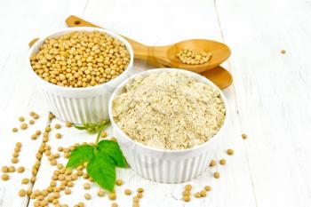 Flour soy and soybeans in two white bowls, spoons and green leaves on the background of light wooden boards
