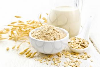 Flour oat in bowl, milk in a jug, oatmeal in spoon, oaten stalks on white wooden board background
