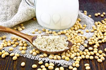 Soy flour in a spoon and milk in a glass jug, soybeans on napkin of burlap on wooden board background
