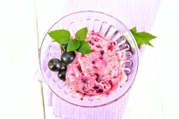 Ice cream with black currant in a glass goblet on a lilac napkin, berries with leaves on a wooden plank background from above