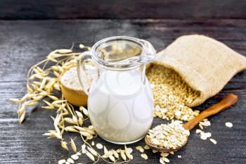 Oat milk in a jug, flour in bowl, oatmeal in a spoon, grain in bag, oaten stalks on the background of dark wooden board